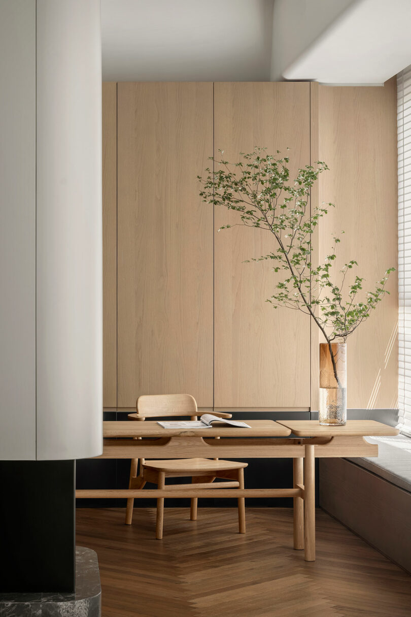 Minimalist dining area with wooden walls and flooring, a wooden table and chair set, and a tall vase with branches. Sunlight filters through a window with blinds.