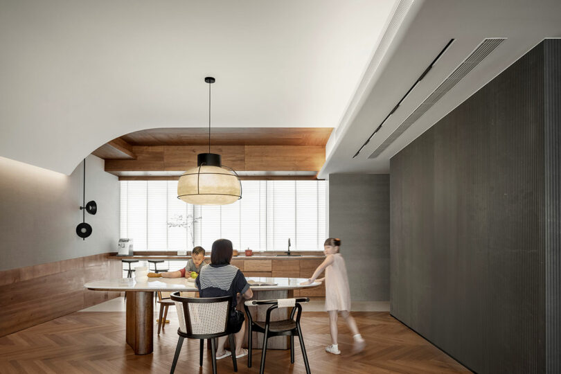 A woman and two children gather around a kitchen island in a modern kitchen with a minimalist design, wood accents, and large windows.