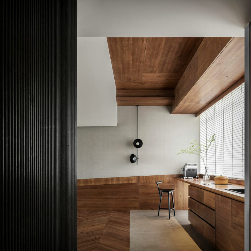 Modern kitchen with wood paneling, minimalist design, a stool at a wooden counter, and large windows with blinds. A small plant and a coffee machine are on the countertop.