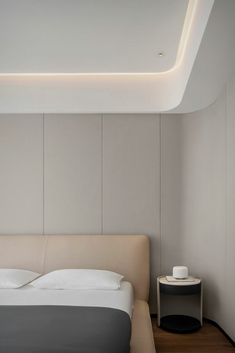 A minimalist bedroom with a bed featuring beige upholstered headboard, white sheets, and black comforter, next to a small round nightstand holding a white cylindrical object, against a gray paneled wall.