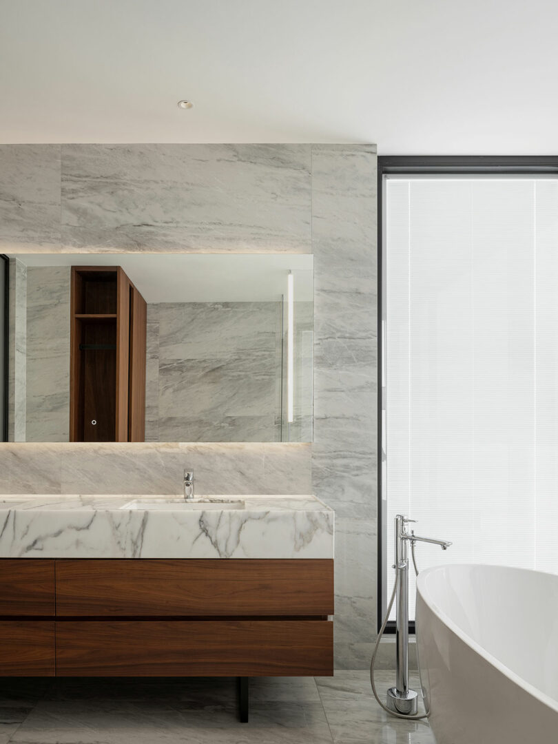 Modern bathroom with marble walls, a large mirror above a wooden vanity, and a white freestanding bathtub near a window with blinds.