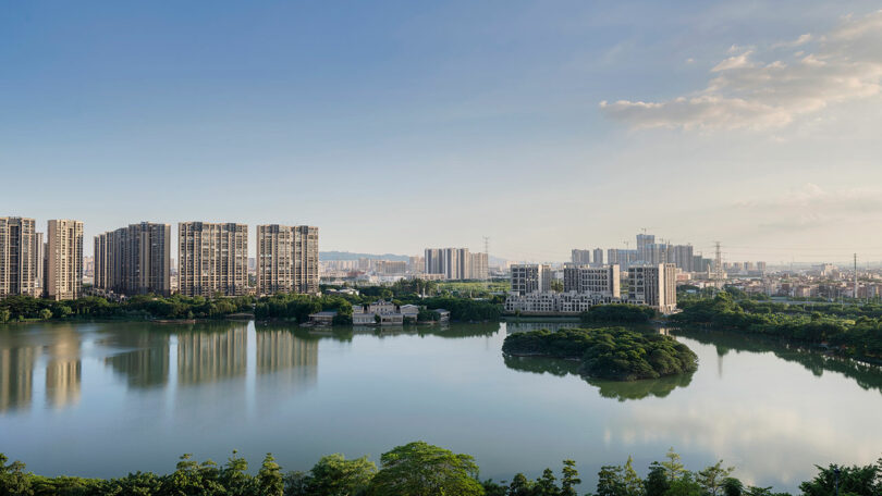 A serene cityscape featuring several high-rise buildings near a calm lake with lush green islands and a tree-lined shore under a clear blue sky.