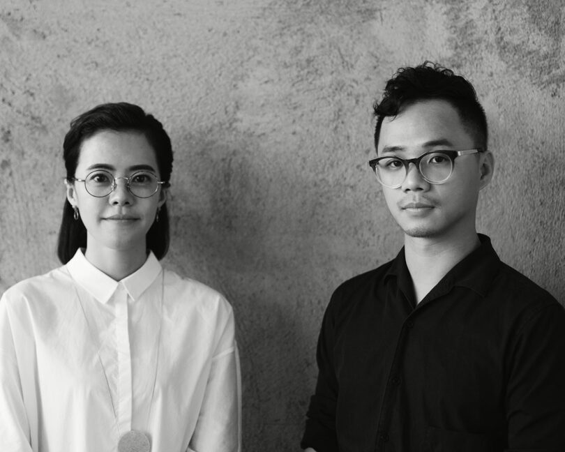 A black and white photo of two individuals with neutral expressions standing against a textured wall. The person on the left wears glasses and a white shirt; the one on the right wears glasses and a dark shirt.