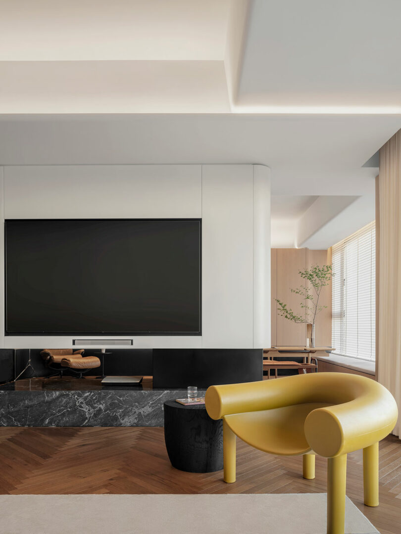 A modern living space featuring a large flat-screen TV mounted on a wall, a yellow contemporary chair, a small side table, and a seating area with a wooden dining table visible in the background.