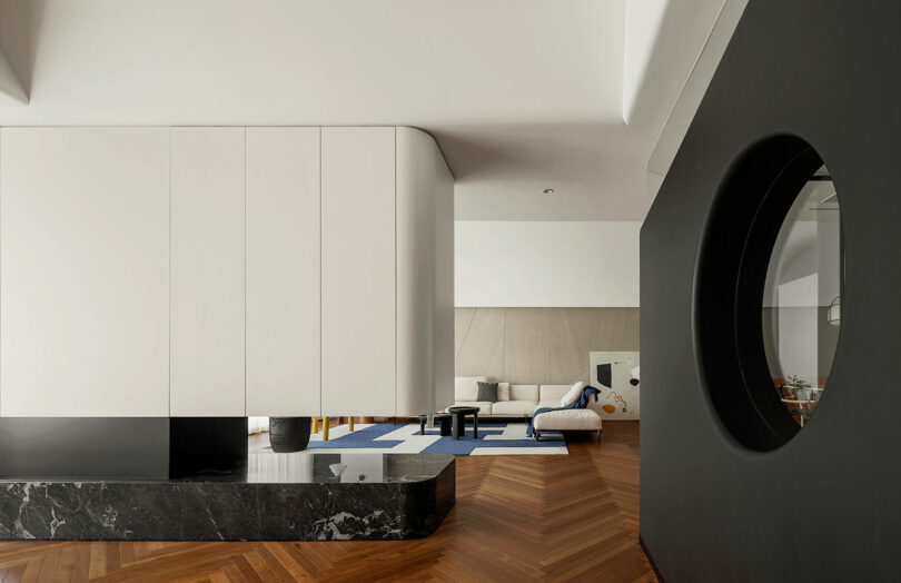 Modern living room with wooden herringbone flooring, a black wall with a circular window, and a white sectional sofa on a blue and beige rug.