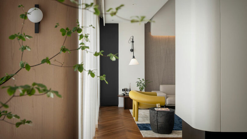 A modern living room with a yellow sofa, white walls, and wooden flooring. A potted plant is in the foreground, and the space is decorated with contemporary lighting fixtures and minimalist furniture.