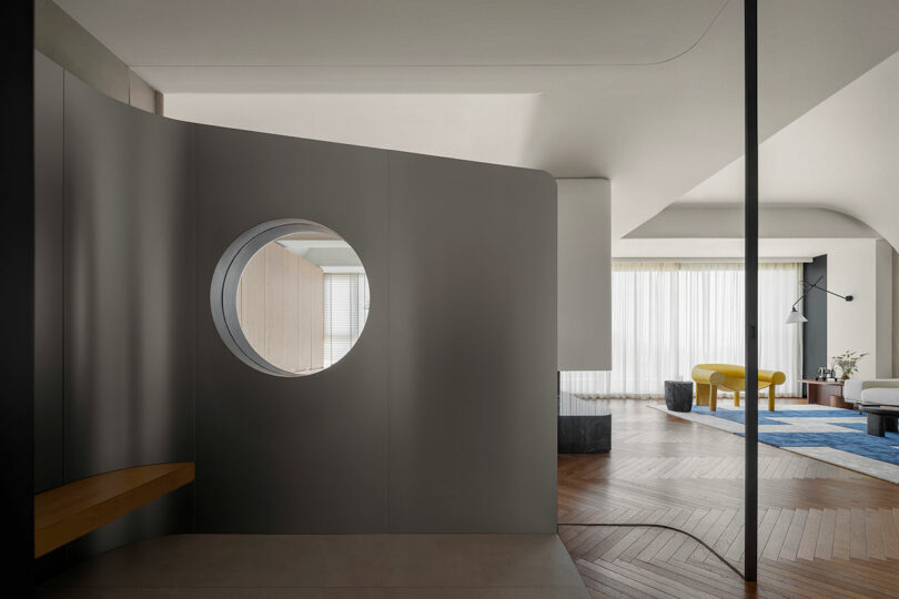 Modern interior featuring a curved wall with a circular window, wooden bench, and view of an open-plan living area with wooden floors, a yellow sofa, a blue rug, and tall white curtains.