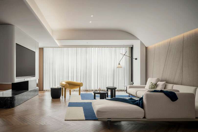 A modern living room featuring a large TV, a white sectional sofa, a yellow armchair, and a geometric rug. The room has wooden flooring and is illuminated by natural light through sheer curtains.