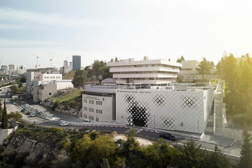 A large, modern building with geometric white tiles on the exterior, situated on a hillside with surrounding trees and nearby cityscape in the background. Several cars are parked along the road.