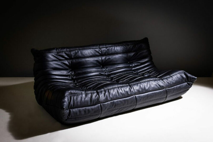 A black leather sofa with a tufted design on seat and backrest, set against a dark background.