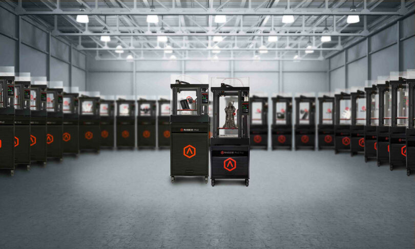 A large industrial room filled with numerous identical 3D printers lined up. Two 3D printers are in the foreground, with one actively printing an object.