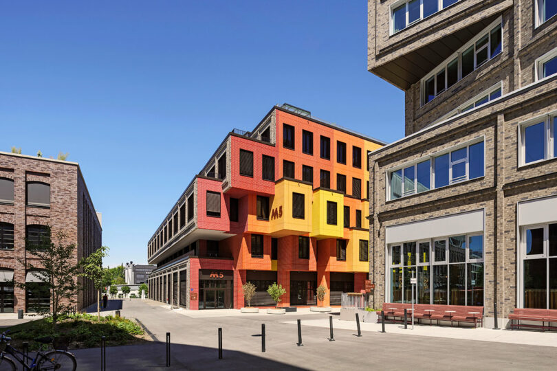 A vibrant, multi-colored building with the letters "M5" designed by Matthias Hollwich stands between two modern, brick structures under a clear blue sky. The street in front is lined with bollards and benches.