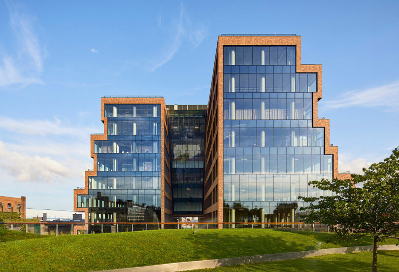 A modern building with a unique tiered brick and glass facade stands under a clear blue sky, surrounded by green lawns and trees.