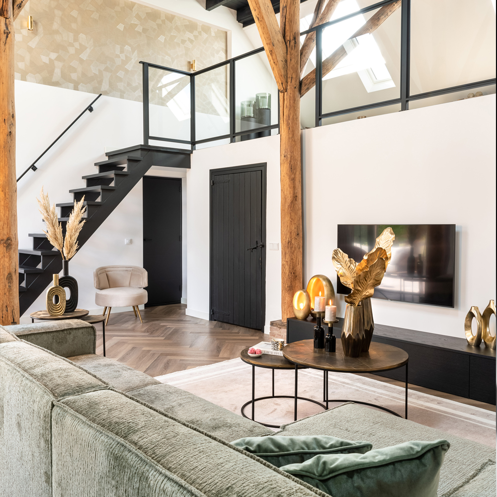 A contemporary living room with a loft and exposed wooden beams. It features a green couch, a wall-mounted TV, a black door, and a staircase with a glass railing. Decor includes gold accents, pampas grass, and a cream armchair paired with the Modern Khaki Fabric Occasional Chair. The floor is designed in an elegant herringbone pattern.