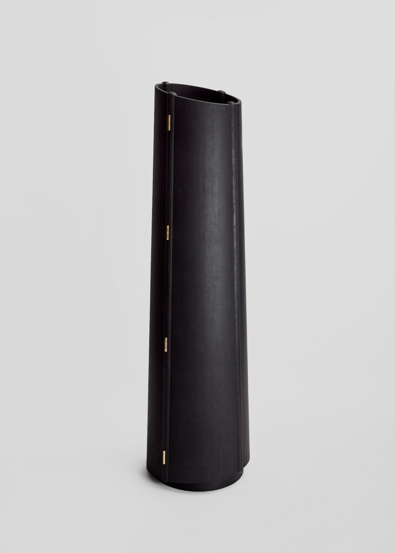 Tall, cylindrical black vase with vertical brass hinges on a light gray background.