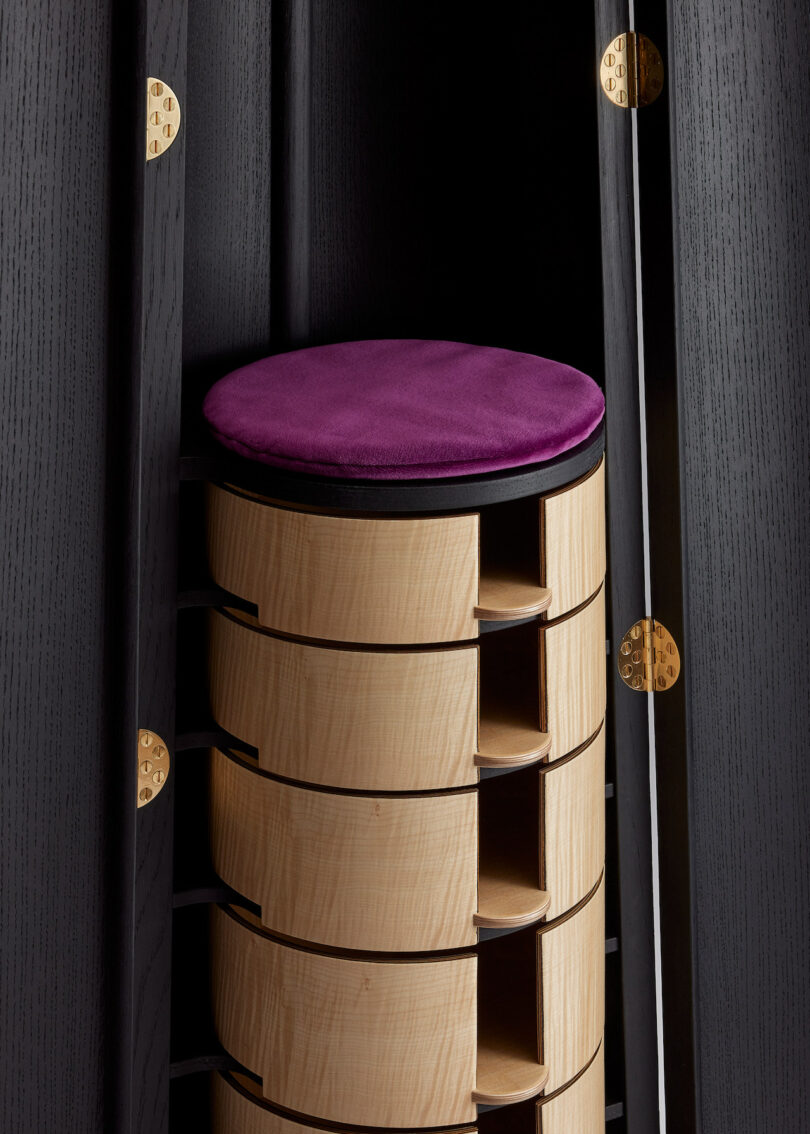 A cylindrical wooden stool with a purple cushion is stored in a tall, black cabinet with gold hinges.