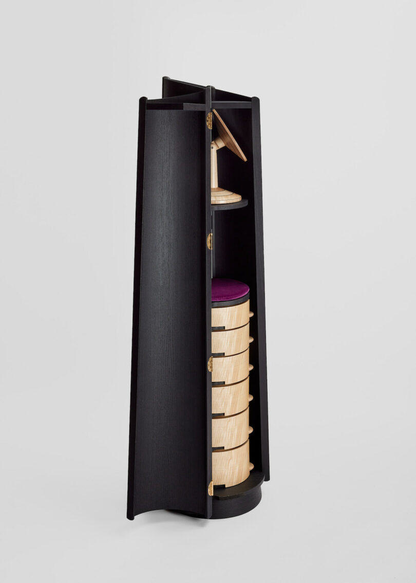 Tall, black cylindrical cabinet with four drawers and an open shelf on top, featuring a wooden accent and a small attached mirror.