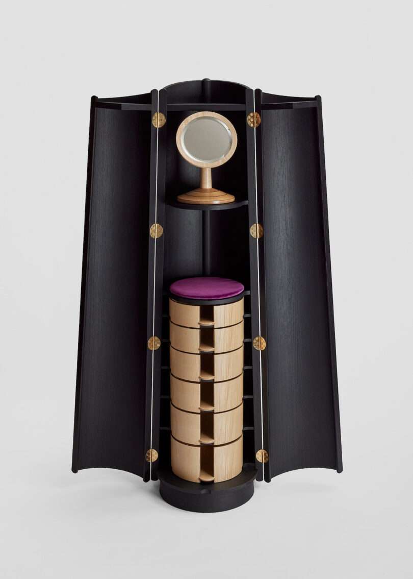 Tall black wooden cabinet with open doors, featuring a round mirror on top and a central stack of circular drawers with a purple cushion.