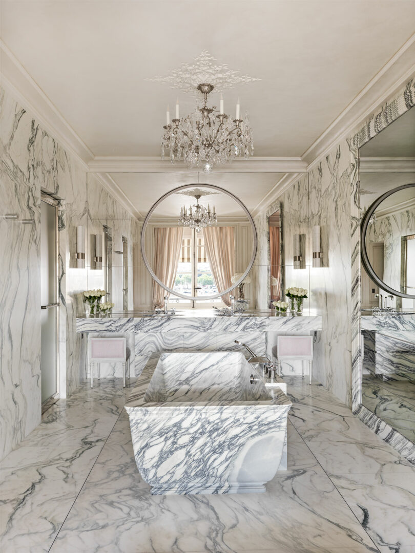 Luxurious marble bathroom with a freestanding tub, large round mirror, chandelier, and two windows with curtains.