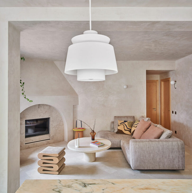 Modern living room with light neutral colors, featuring a gray sectional sofa, round coffee table, distinctive stacked stool, and a contemporary pendant light. A fireplace is built into the wall.