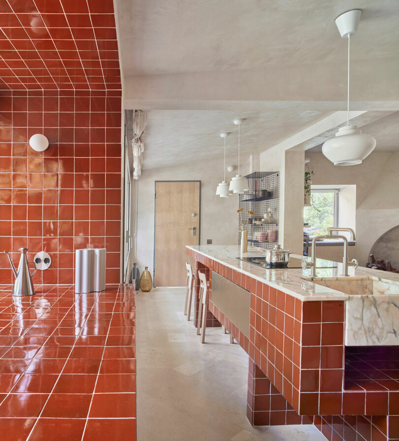 A modern kitchen with red tile countertops and walls, marble island, wooden stools, and pendant lights. The space includes a stainless steel trash can and various kitchen appliances. A door is in the background.