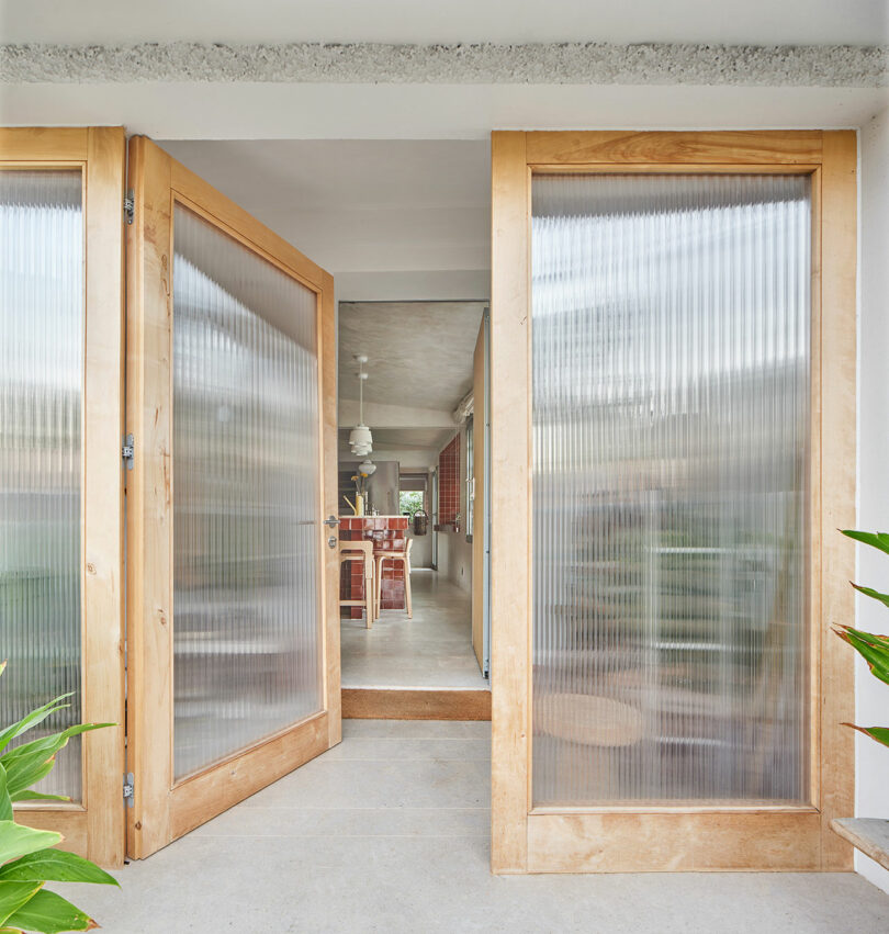 A modern entryway with large wooden-framed glass doors, giving a view into a bright, minimalistic interior with wooden furniture and plants.