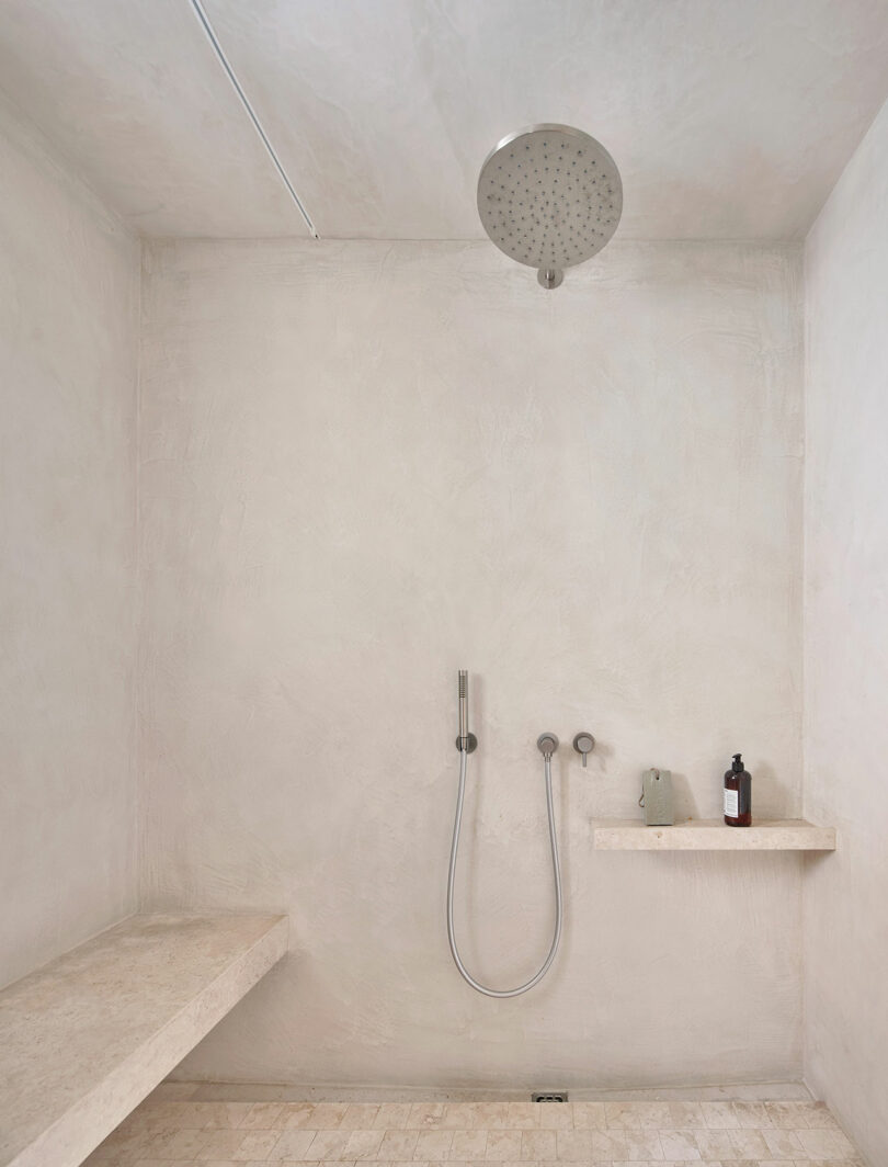 Minimalist bathroom with a rain shower head, handheld shower, a built-in bench, and a small shelf holding a soap dispenser and a green container. Walls and floor are in light beige tones.