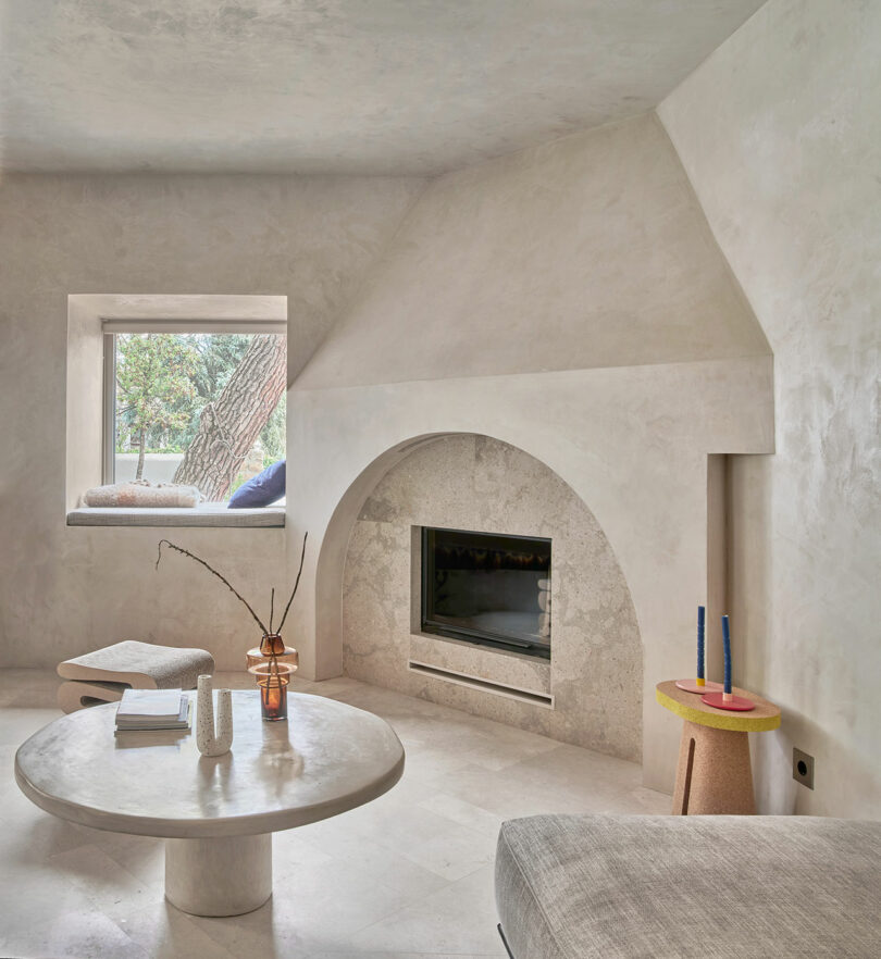 Minimalist living room with a fireplace, round coffee table, books, and vase with branches. Window seat with cushions on the left, candlesticks on a side table to the right, and greenery visible outside.