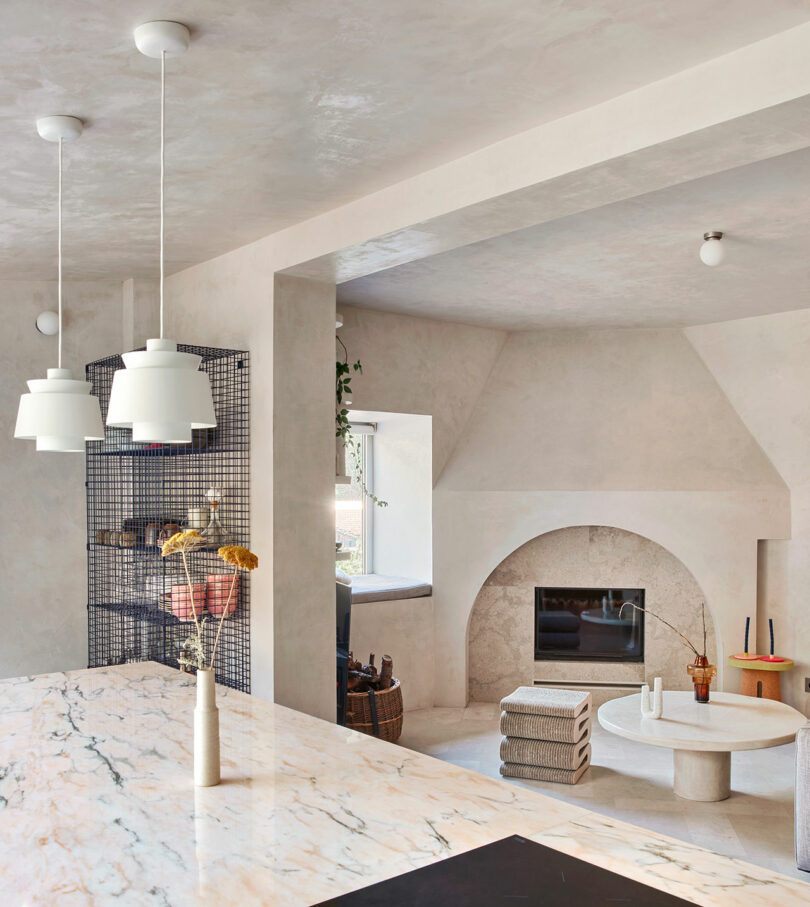 Modern living area with a marbled kitchen counter, hanging white pendant lights, and a cozy seating nook by a stone fireplace. The space includes a round coffee table with decorative items.