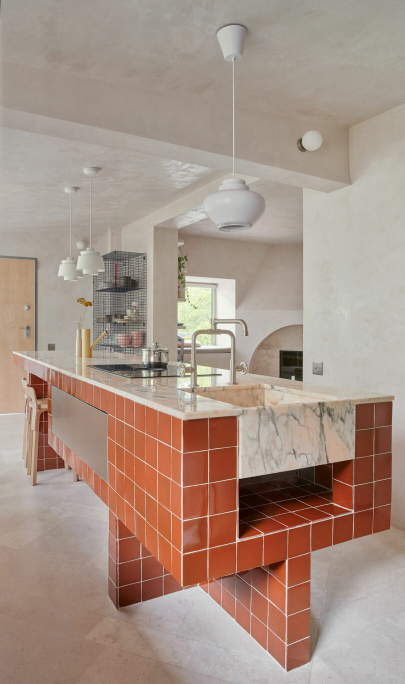 A modern kitchen features a long red tile island with a marble countertop. Pendant lights hang above, and minimalist decor includes jars and a potted plant. A wire shelf unit is visible in the background.