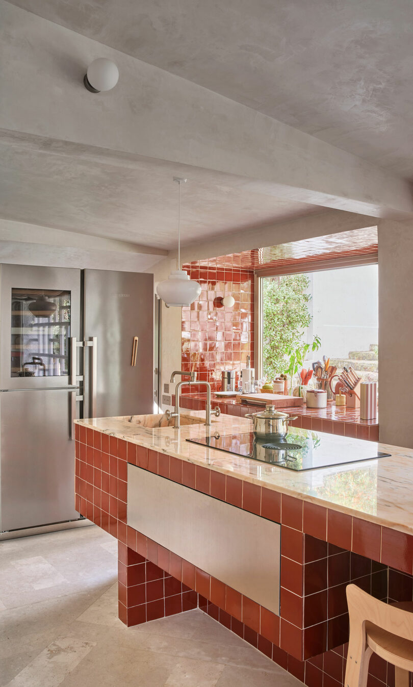 Modern kitchen with red tiled island, stainless steel appliances, large window, and various cooking utensils. Neutral decor with natural light illuminating the space.