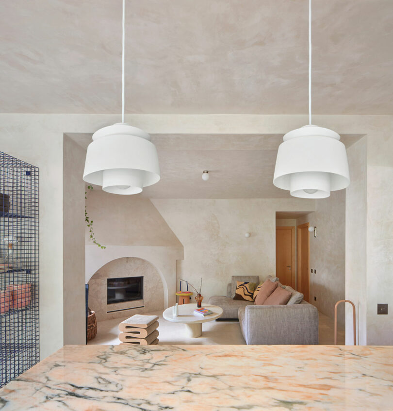 A modern living room with a stone fireplace, gray sofa, and white coffee table. Two white pendant lights hang above a marble countertop in the foreground.