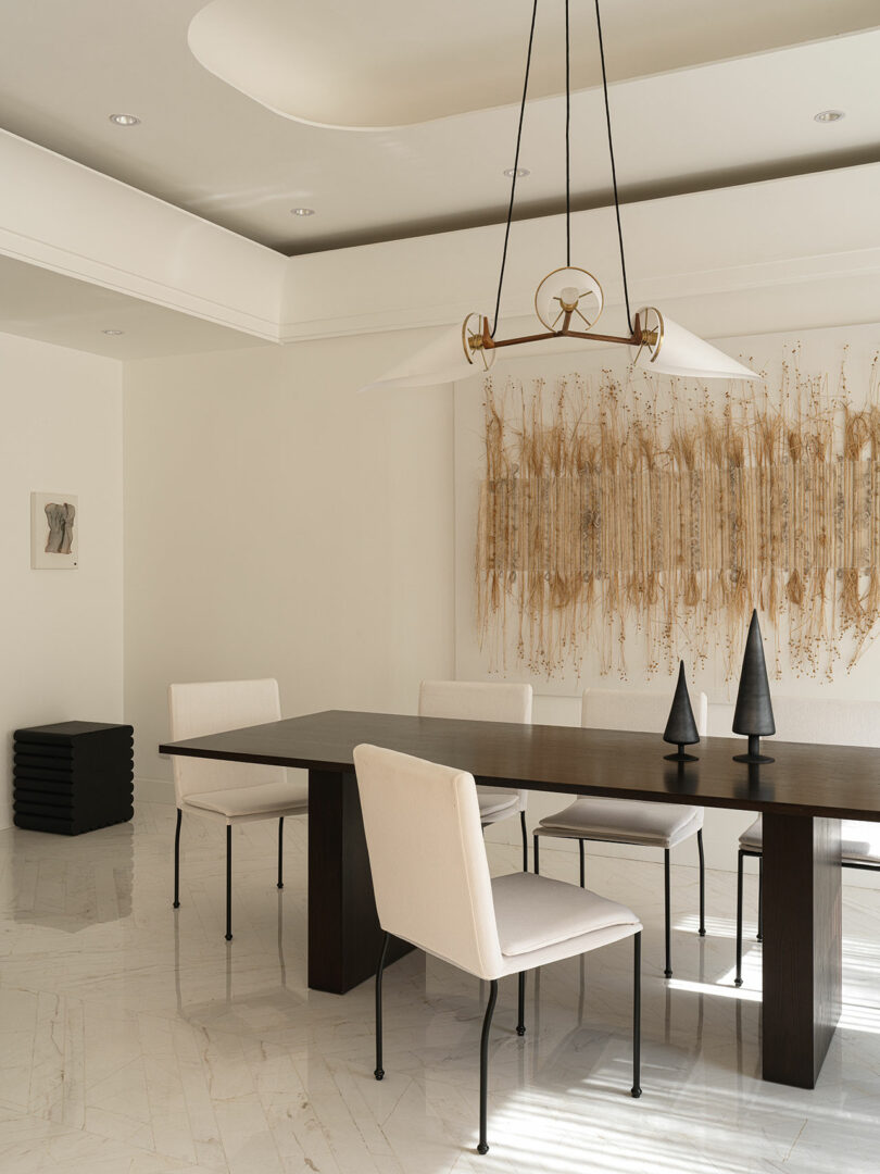 Minimalist dining room with a dark wooden table, four white chairs, a modern pendant light, wall art featuring natural elements by Marina Braginskaya, and a stack of black stools in the corner.