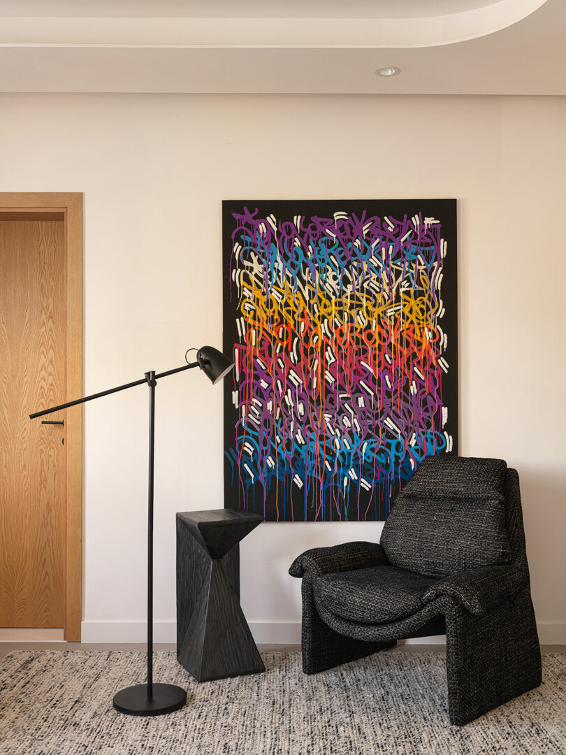 Room corner with abstract wall art by Marina Braginskaya, black standing lamp, black textured chair, and small black side table. White wall, wooden door on the left, and patterned rug on the floor.