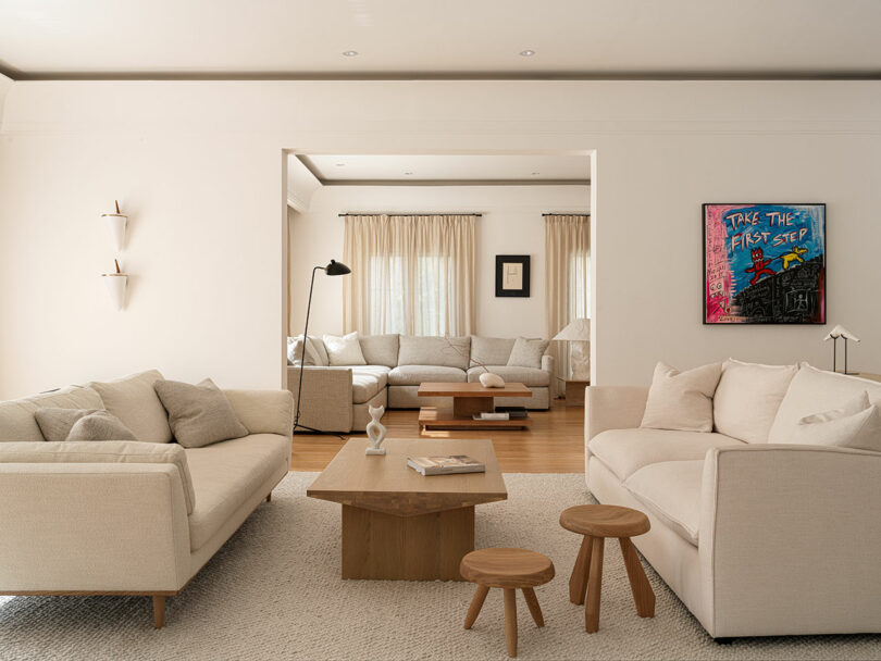 Minimalist living room featuring light-colored sofas, a wooden coffee table, and stools. Artwork by Marina Braginskaya with "TAKE THE FIRST STEP" text adorns the wall. The space boasts a neutral color palette and offers a view into an adjoining room.