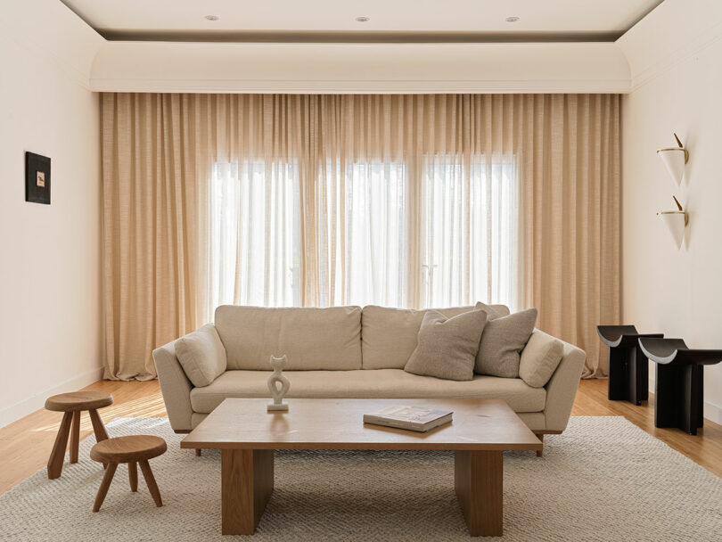 A neutral-toned living room features a beige sofa, wooden coffee table, small side tables, and sheer beige curtains. Minimalist decor includes a small wall shelf and a piece of artwork by Marina Braginskaya.