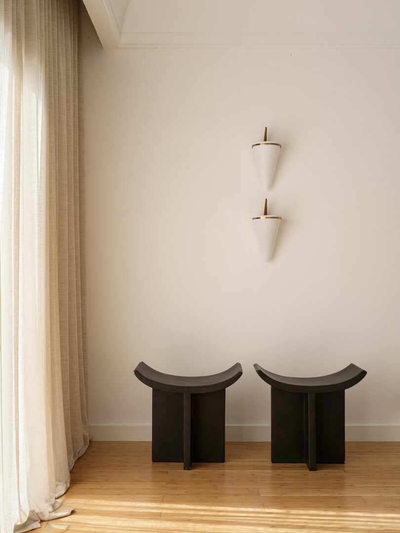 A minimalist interior featuring two black curved stools, a wooden floor, sheer curtains, and two cone-shaped wall sconces installed vertically on a plain white wall—reminiscent of the refined elegance often seen in Marina Braginskaya's designs.