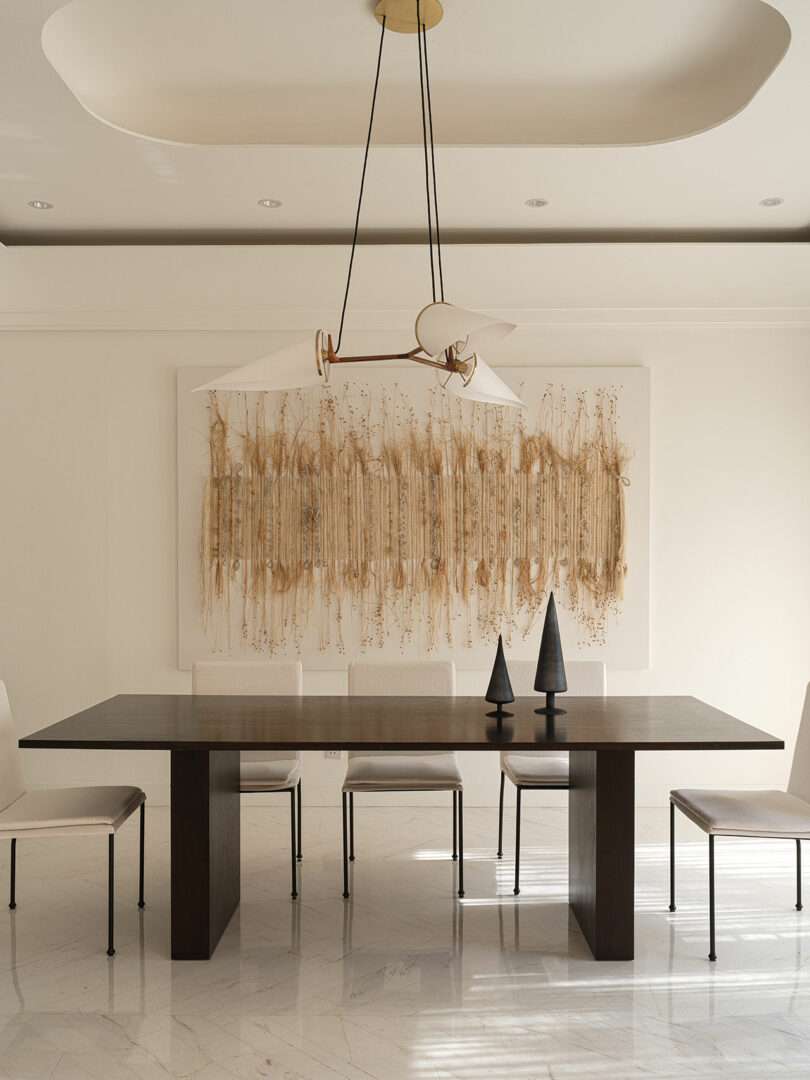 A minimalist dining room features a dark wooden table, four white chairs, abstract wall art with organic textures reminiscent of Marina Braginskaya's style, and a modern light fixture hanging from the ceiling.