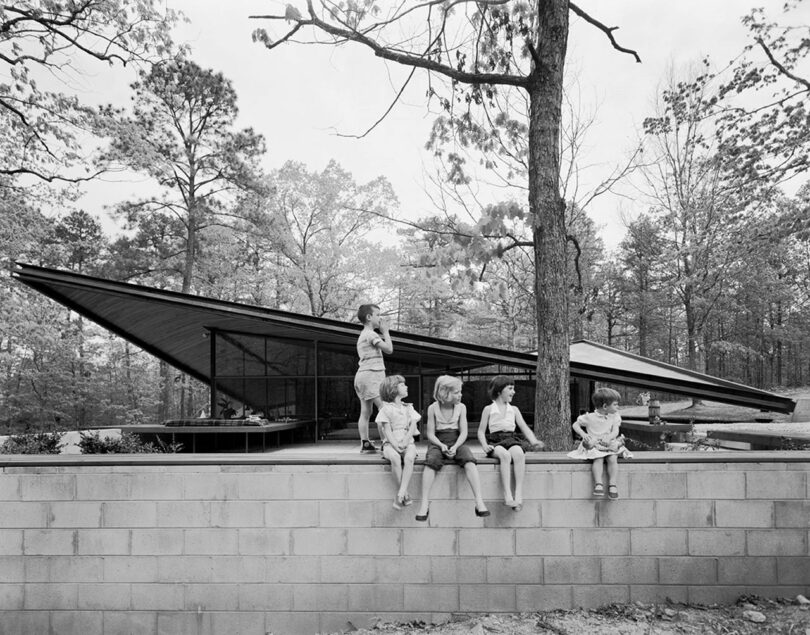 Five children, sit on a low stone wall in front of a modern, glass-walled house surrounded by trees.