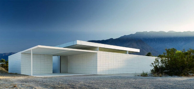 A modern minimalist white building with a flat roof set against a backdrop of desert terrain and mountains under a clear sky.