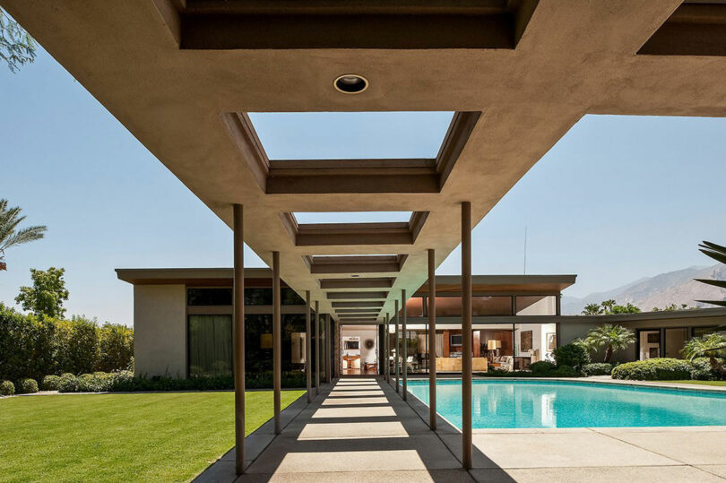 Modern house exterior with a long walkway, rectangular overhead cutouts, green lawn, and a swimming pool. The house features large windows and is surrounded by trees and distant mountains.