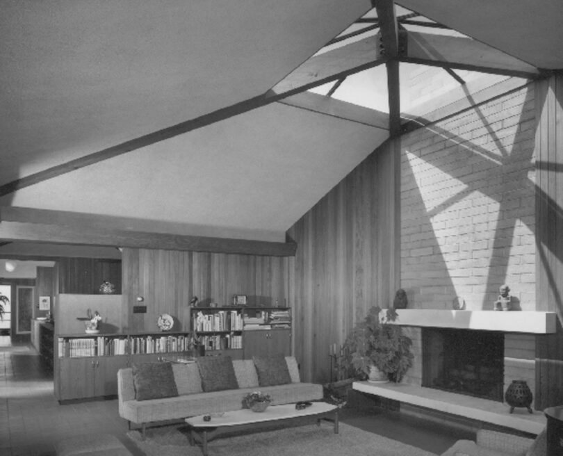 A modern living room with wooden panel walls, a high ceiling with exposed beams, a fireplace, bookshelves, and comfortable seating. Skylight casts shadows on the brick wall above the fireplace.