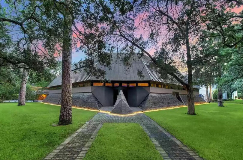 A large, modern wooden house with a sloped roof, surrounded by trees and grass, with a brick pathway leading to the entrance under a sunset sky.