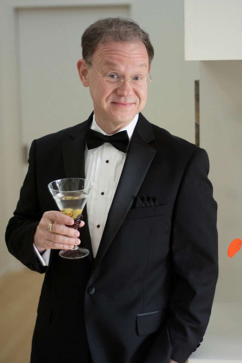 George Smart, wearing a black tuxedo and holding a cocktail glass, stands indoors, smiling at the camera.