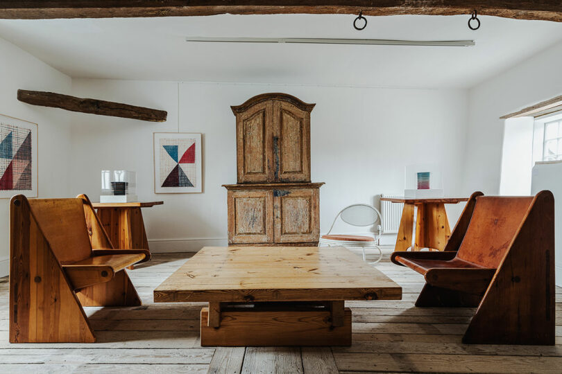 A room with wooden furniture: two benches, a table, and a cabinet. White walls showcase geometric artwork, reminiscent of Richard Parr's style. A wooden beam and a large window are visible.