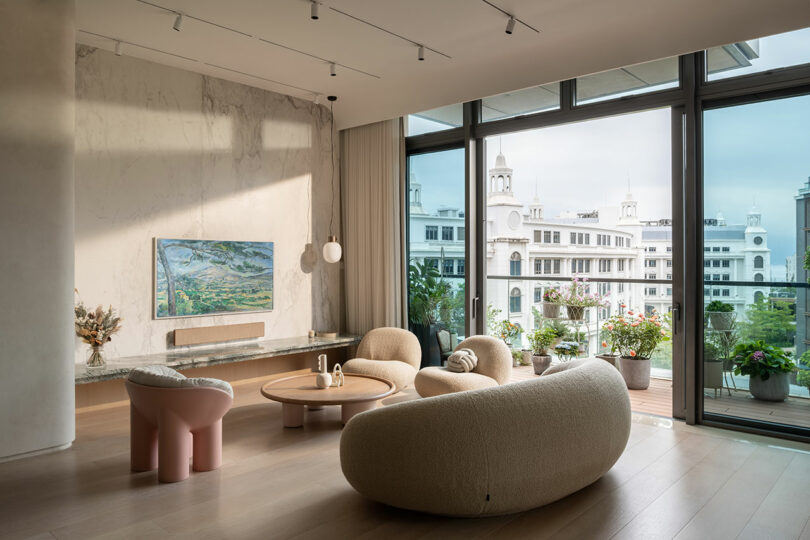 Spacious modern living room with large windows, curved furniture, wall-mounted TV, abstract art, and a balcony with potted plants overlooking a cityscape with domed buildings.