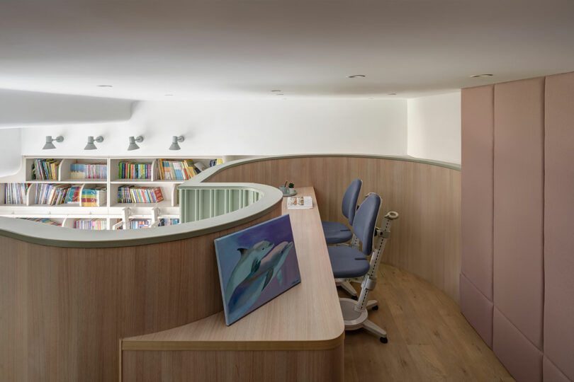 Modern library interior with wooden curved walls, a desk with a dolphin painting, two blue chairs, and bookshelves in the background.