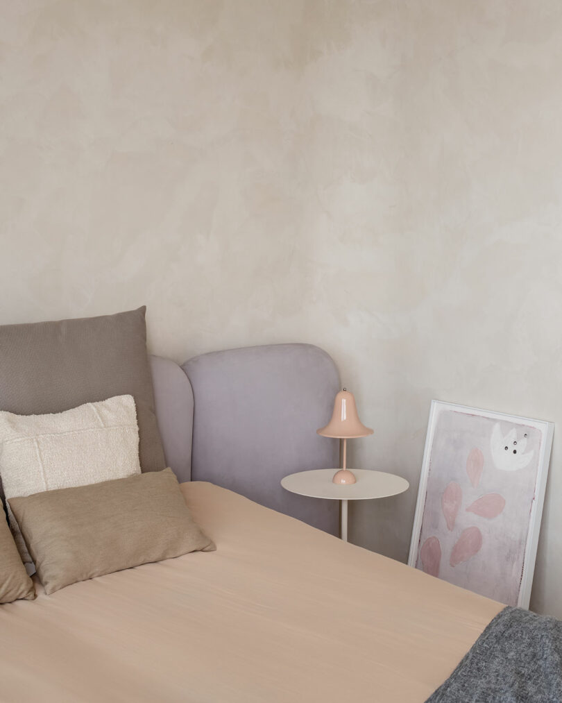 A neutral-toned bedroom with a beige bed, gray and beige pillows, a small side table with a pink lamp, and a framed picture with a cat illustration leaning against the wall.