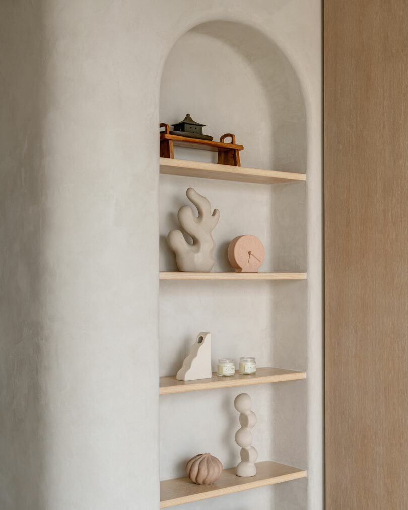 A recessed shelf with four levels, holding minimalist decor items including sculptures, candles, a clock, and a model house. The shelf is built into a light-colored wall with an arched top.