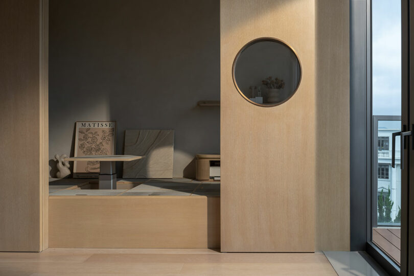 Minimalist interior featuring a wooden platform with art pieces and a circular window. Natural light from the open glass door brightens the space.